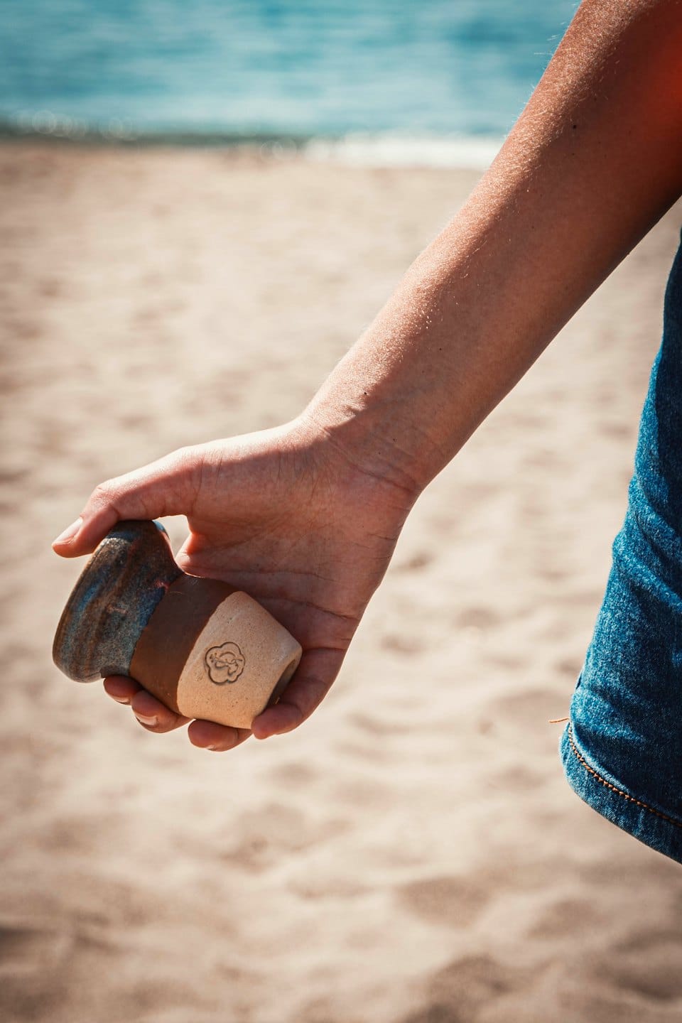 A person holds a handmade ceramic mug near the beach.