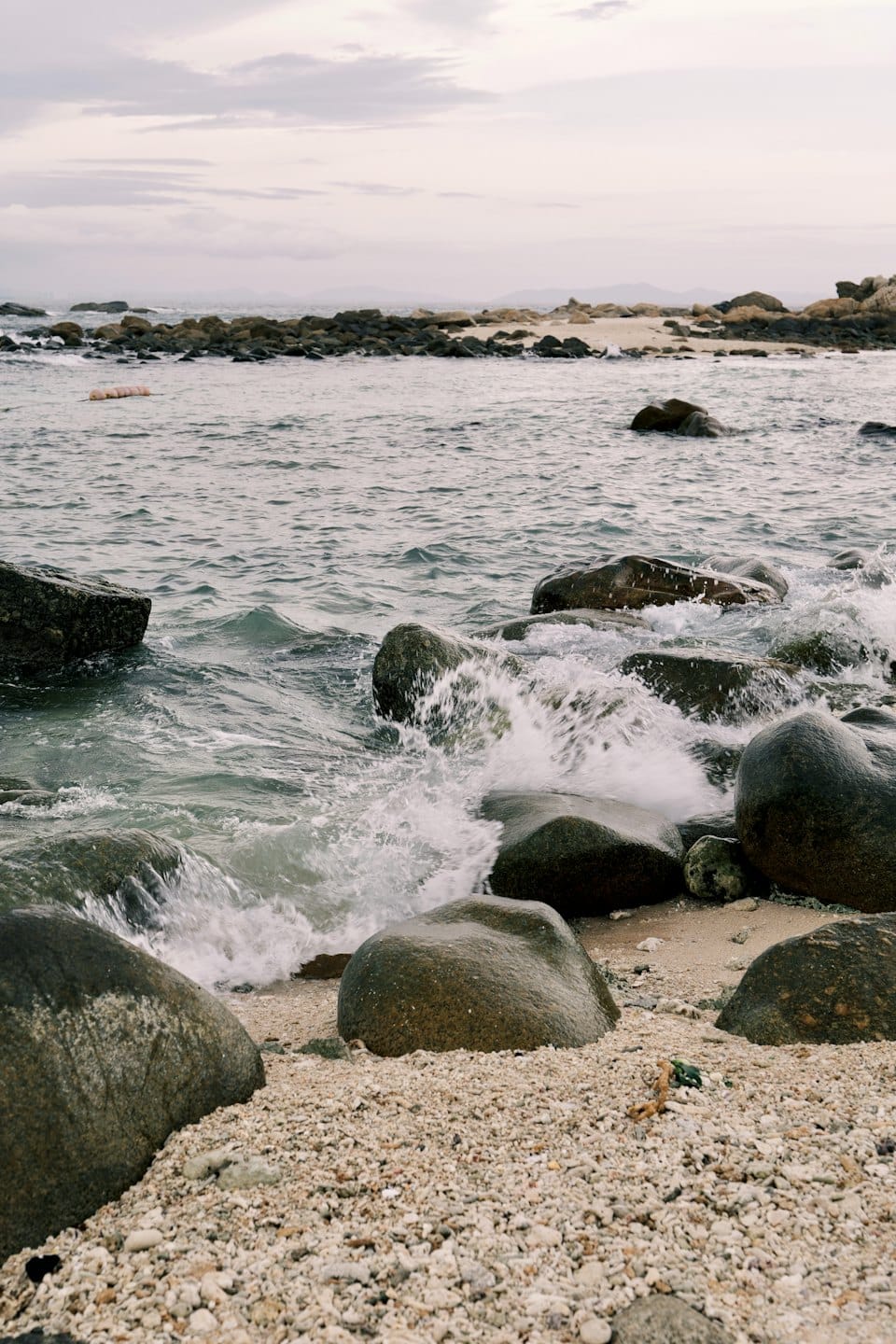 Waves crash against dark rocks on a pebbly beach.